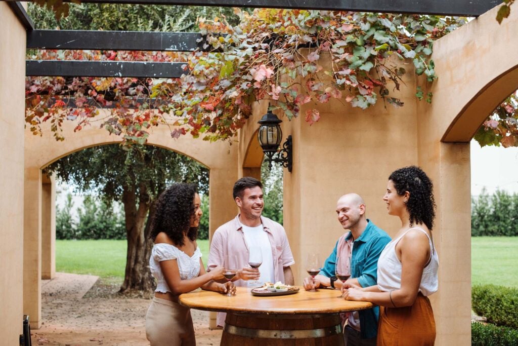 Four friends smiling and enjoying red wine and appetizers around a wooden barrel table in a rustic outdoor courtyard.