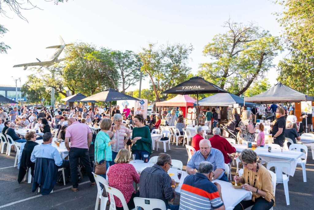 A crowded outdoor food festival or community event with people seated at long white tables under umbrellas and tents