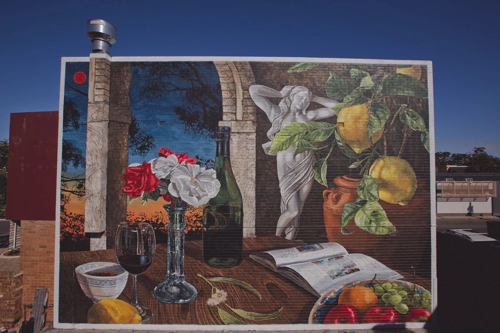 A large, intricate outdoor mural on a brick wall featuring a classical still life with a wine bottle