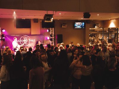 A crowded indoor venue with people watching a live band perform on a stage illuminated by pink lights.