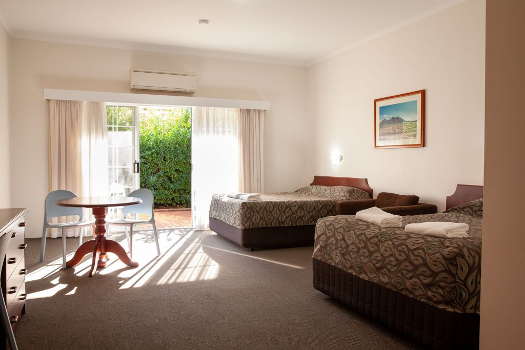 A sunlit hotel room featuring two twin beds with patterned brown bedspreads