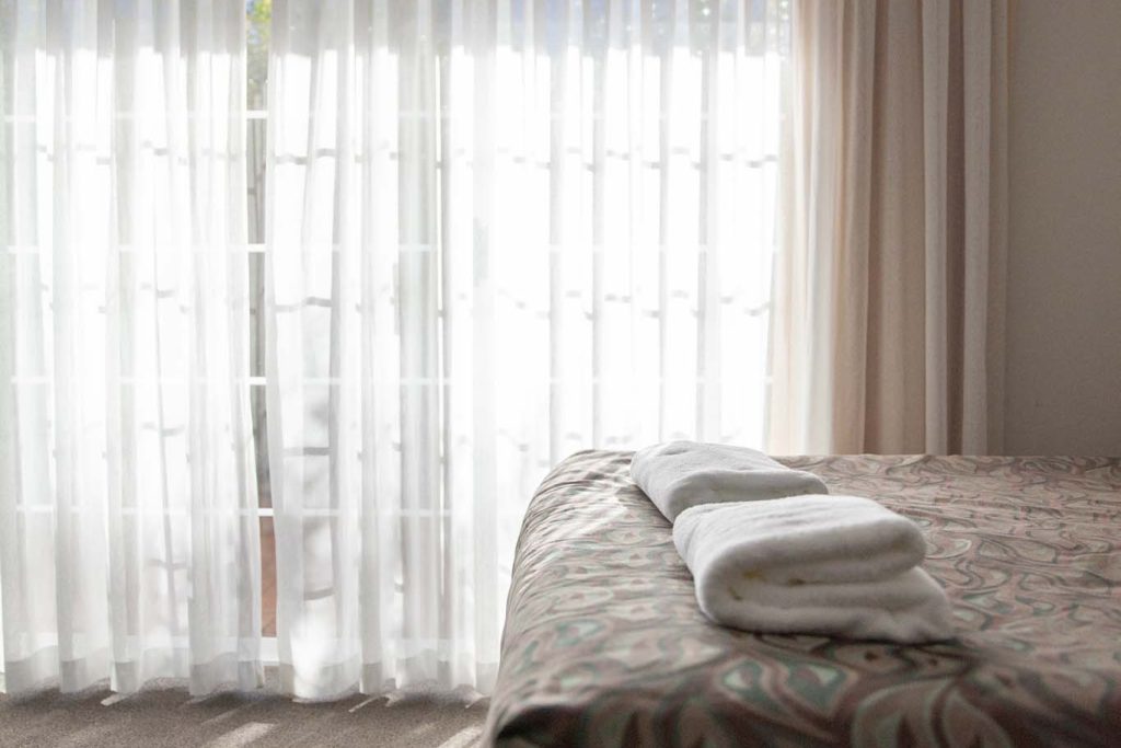The Kidman Wayside Inn room featuring a bed with a patterned bedspread and folded white towels, positioned in front of a large window with sheer white curtains.
