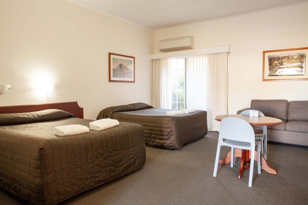 The Kidman Wayside Inn room with two large beds featuring dark brown quilted covers and folded white towels, a small dining table with chairs, and a grey sofa.