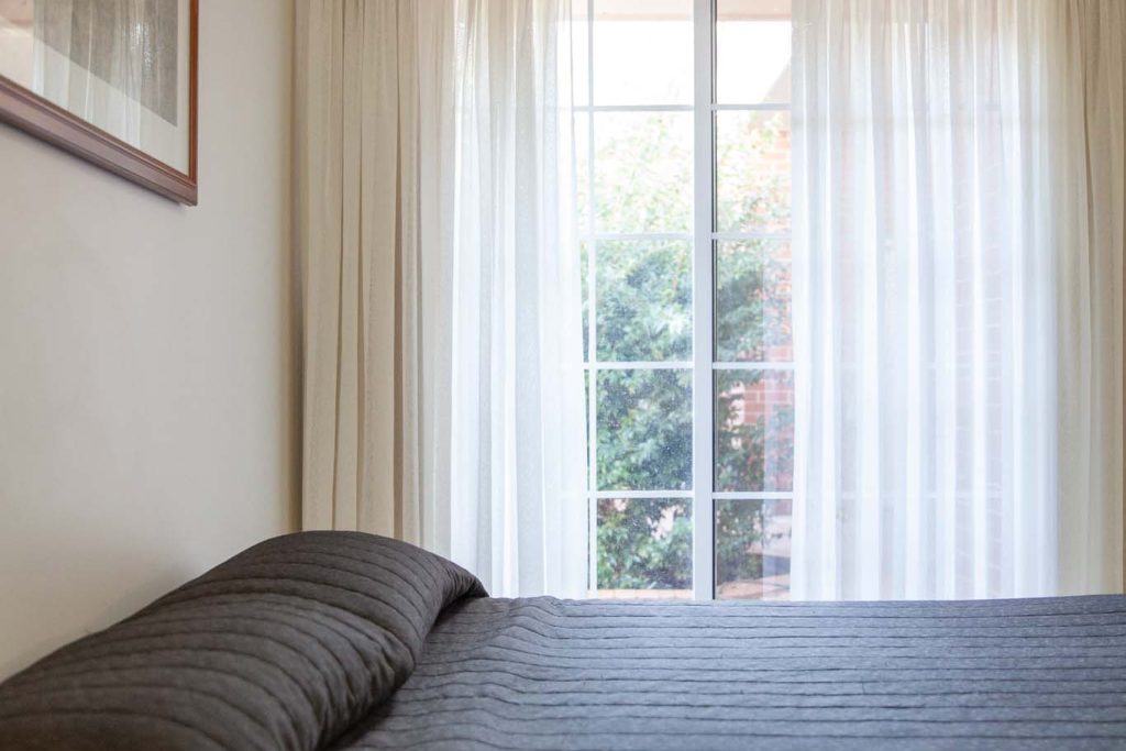 The Kidman Wayside Inn bedroom featuring a dark grey quilted bed in the foreground, white sheer curtains over a window, and a wall-mounted air conditioner.