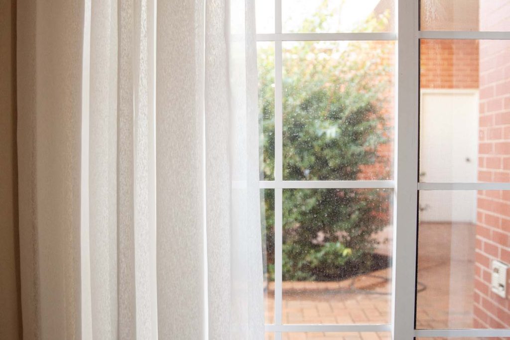 The Kidman Wayside Inn window with white sheer curtains and a white grid frame, revealing an outdoor courtyard.