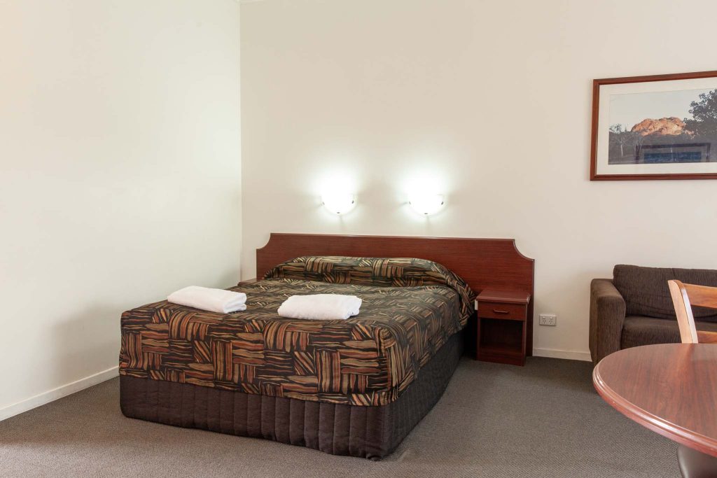 A brightly lit motel room featuring a large bed with a patterned brown bedspread and a small wooden nightstand