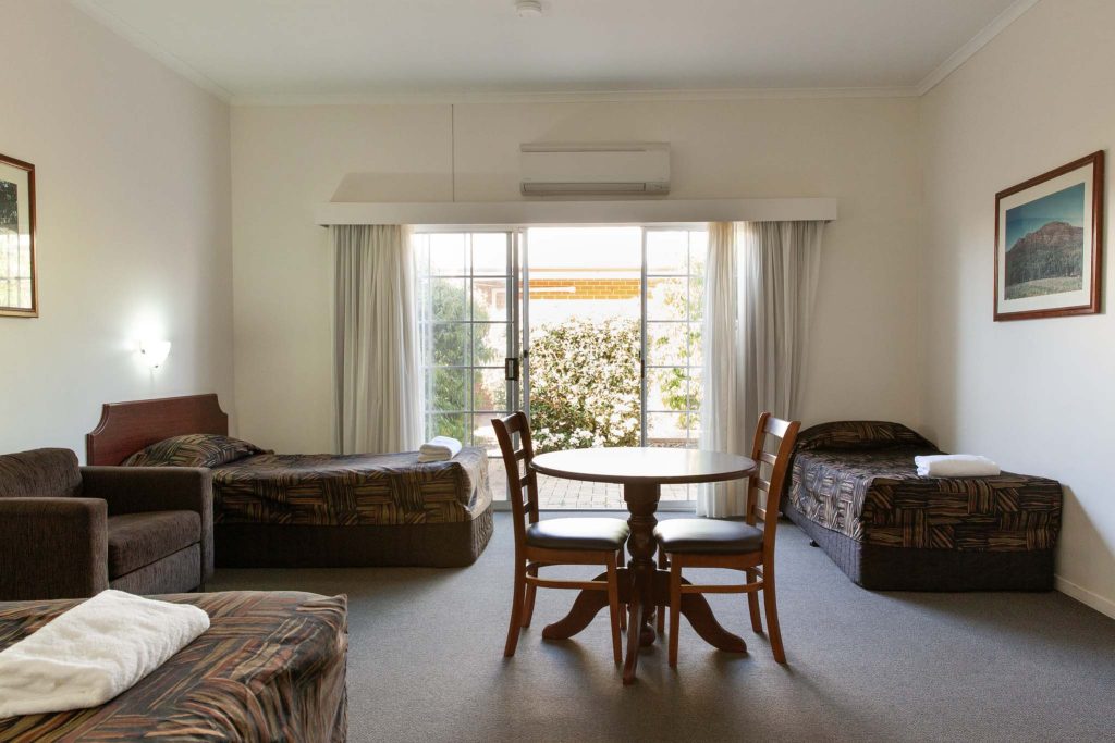 A spacious hotel room viewed from the centre, showing three single beds with matching patterned brown covers.