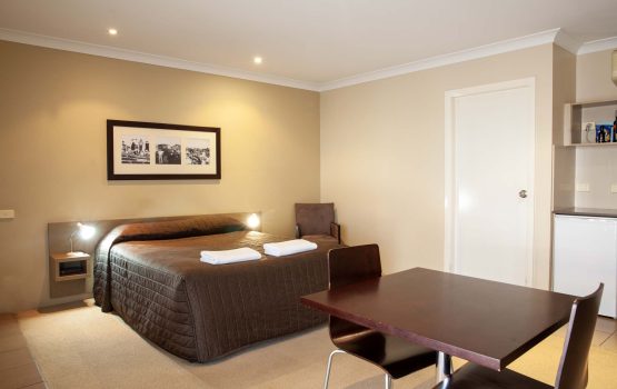 The Kidman Wayside Inn beige-toned room featuring a queen bed with a brown quilted cover two bedside lamps, and a framed triptych of vintage photos on the wall.
