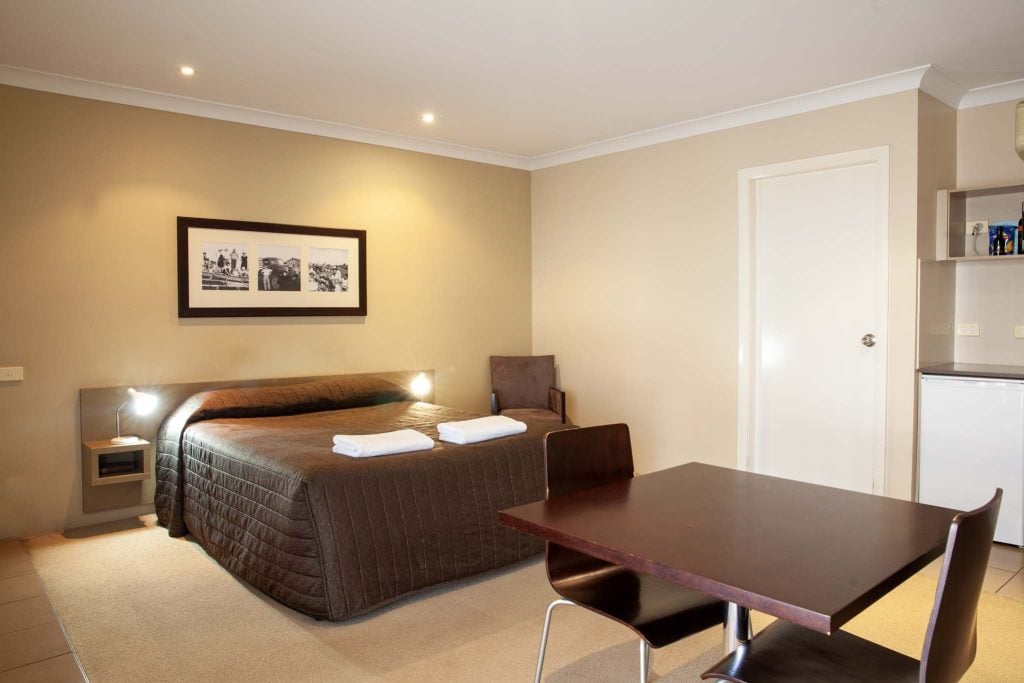 The Kidman Wayside Inn beige-toned room featuring a queen bed with a brown quilted cover two bedside lamps, and a framed triptych of vintage photos on the wall.